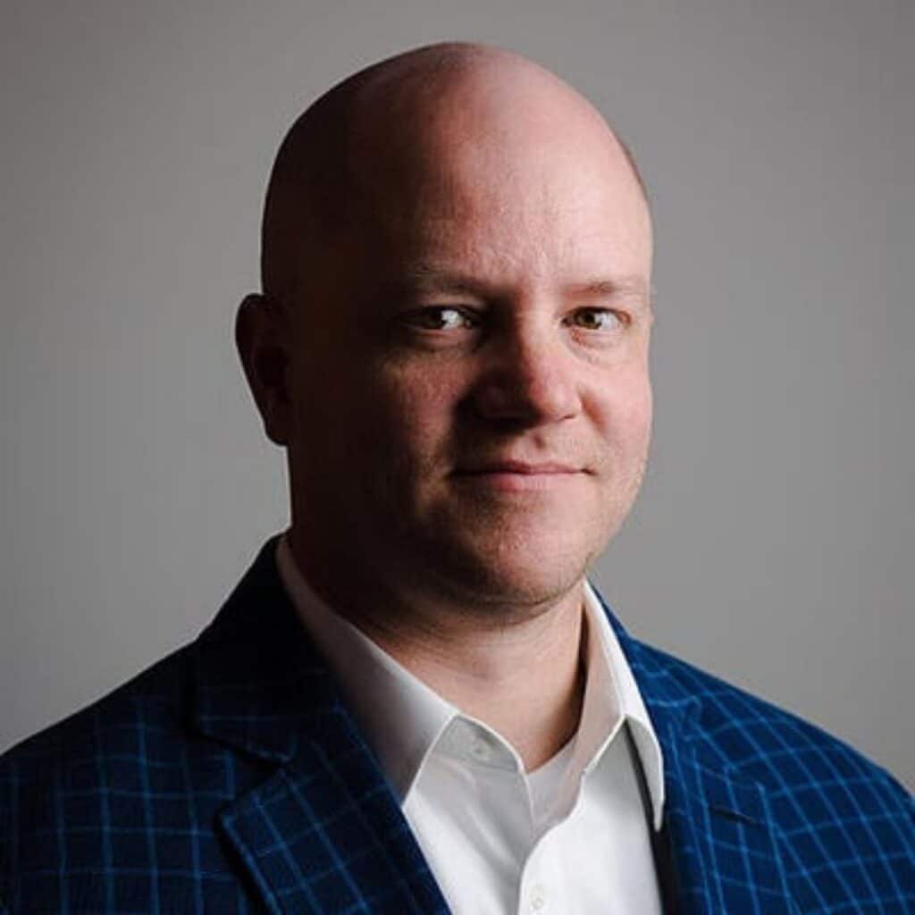 A bald man wearing a blue checkered suit jacket and white dress shirt stands in front of a plain gray background, looking slightly to the side with a neutral expression. C3 Solutions