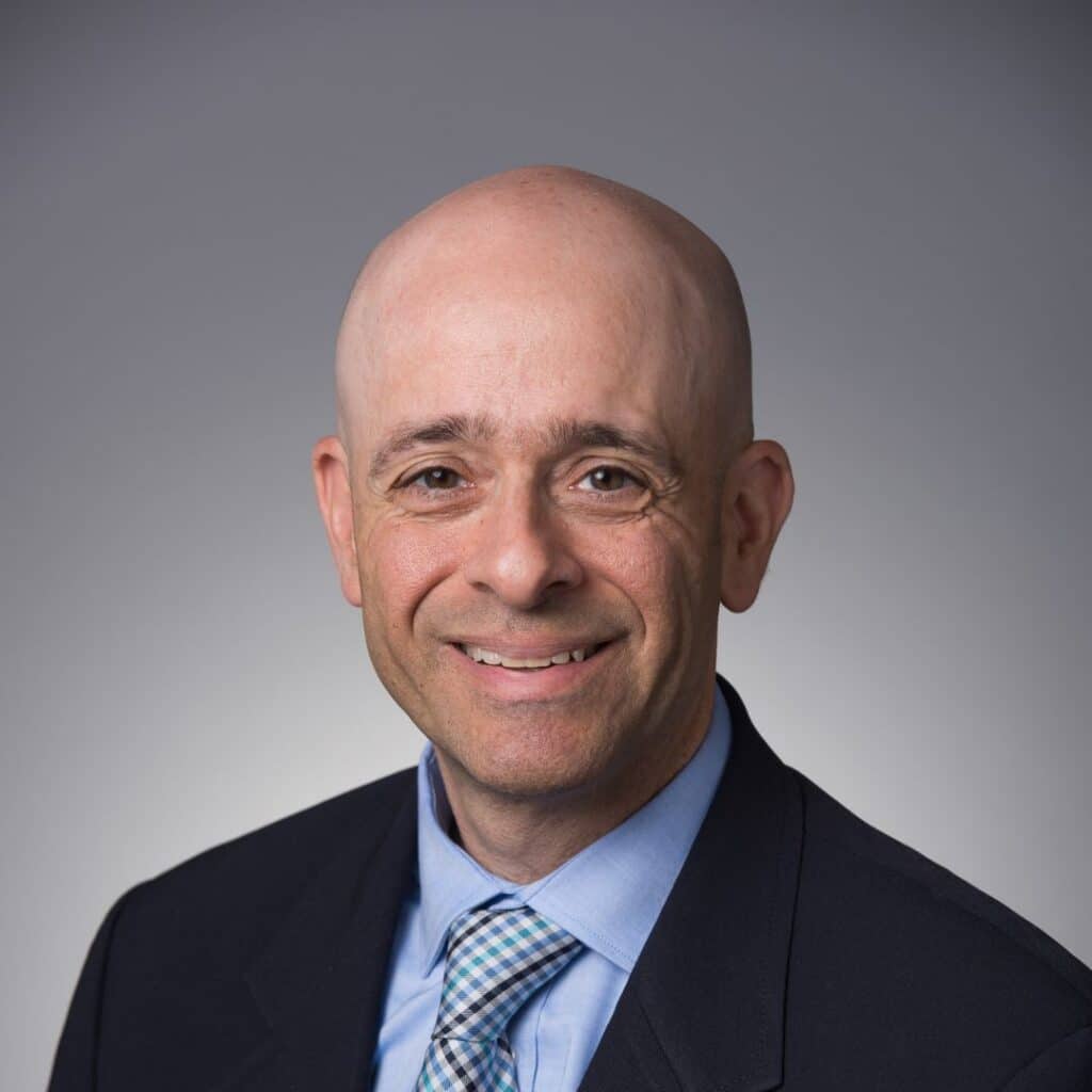 A smiling bald man wearing a dark suit, light blue shirt, and plaid tie poses in front of a plain gray background. C3 Solutions