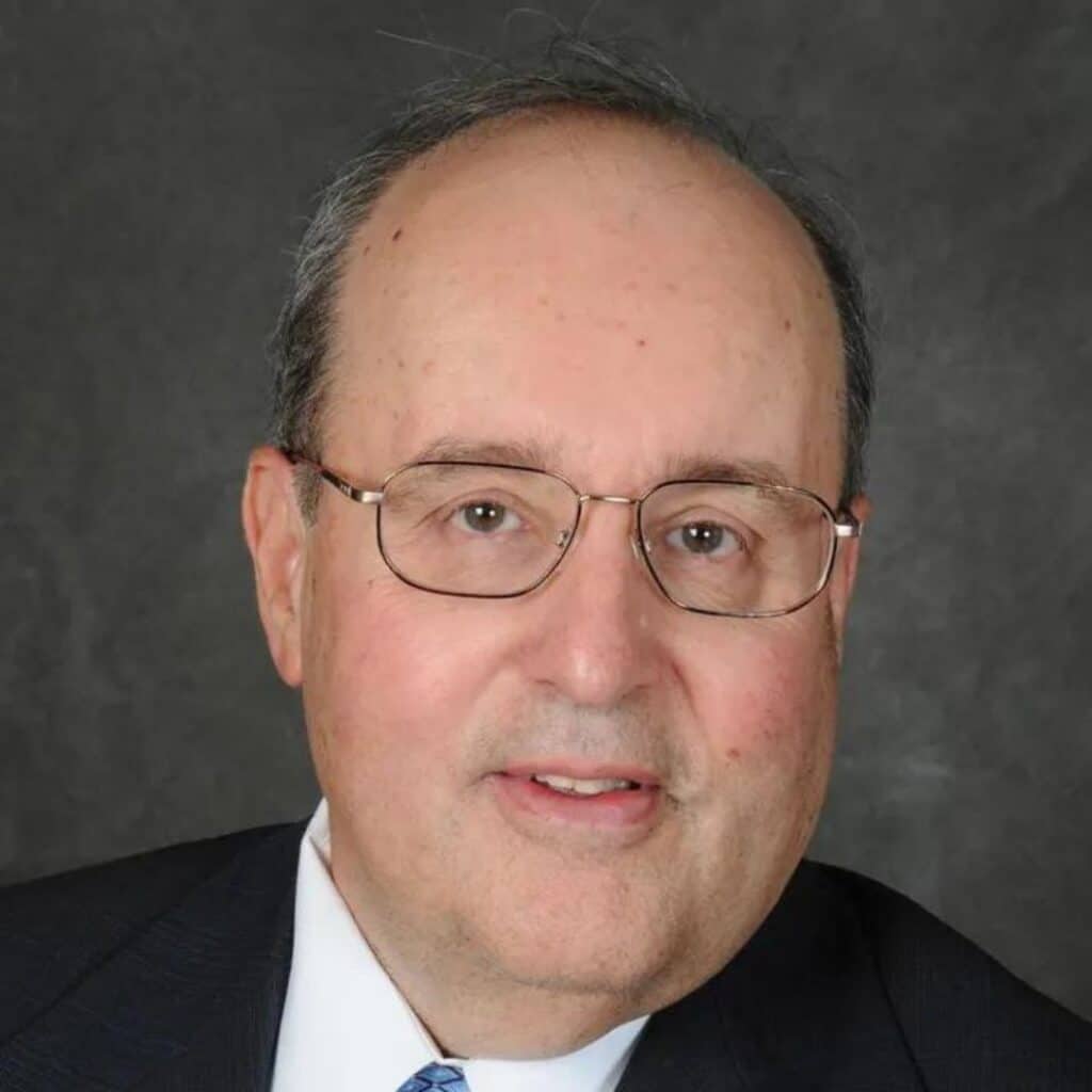 A middle-aged man with glasses and thinning hair, wearing a dark suit jacket, white shirt, and blue tie, poses in front of a gray background. C3 Solutions