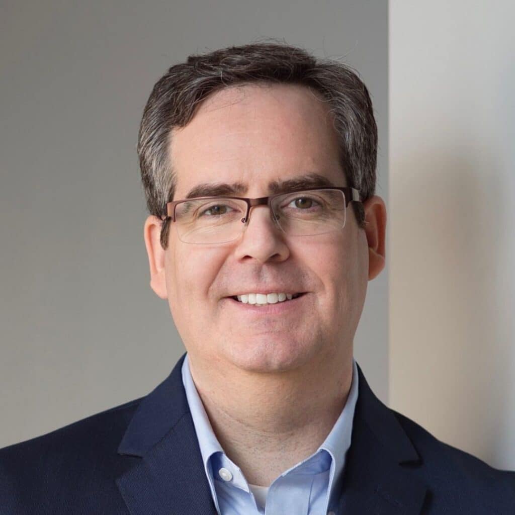 A man with short dark hair, glasses, and a light blue shirt under a dark blazer smiles warmly while standing indoors against a neutral background. C3 Solutions