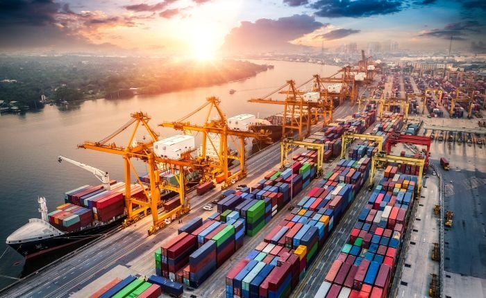 A busy shipping port at sunset, with large cranes loading colorful shipping containers onto cargo ships beside a wide river, and rows of stacked containers—some incurring demurrage-charges—lining the dock. C3 Solutions