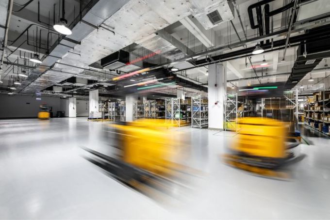 Un entrepôt spacieux et moderne, avec des rayonnages remplis de cartons et plusieurs robots automatisés jaunes — équipés de « outils d’automatisation » — qui se déplacent rapidement sur le sol poli, créant un effet de flou de mouvement. Le plafond est orné de tuyaux apparents et d’un éclairage industriel. C3 Solutions