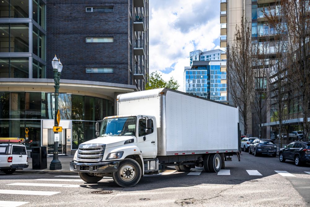 A white box truck is turning at an intersection in a modern city, demonstrating the importance of blind shipment logistics in trucking, with tall buildings, trees, and several parked cars visible on a sunny day. C3 Solutions