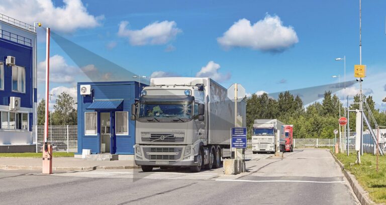 Three large trucks are lined up at a security checkpoint with a blue guardhouse. A raised barrier and various signs are visible under a clear blue sky with clouds. Trees and a fence are in the background. C3 Solutions