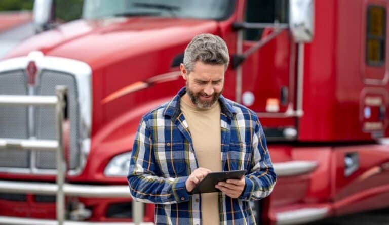 Un homme vêtu d'une chemise à carreaux se tient devant un semi-remorque rouge, souriant tandis qu'il utilise une tablette en extérieur — illustrant ainsi comment les chauffeurs peuvent facilement gérer la planification des quais et optimiser leurs opérations grâce aux technologies modernes. C3 Solutions