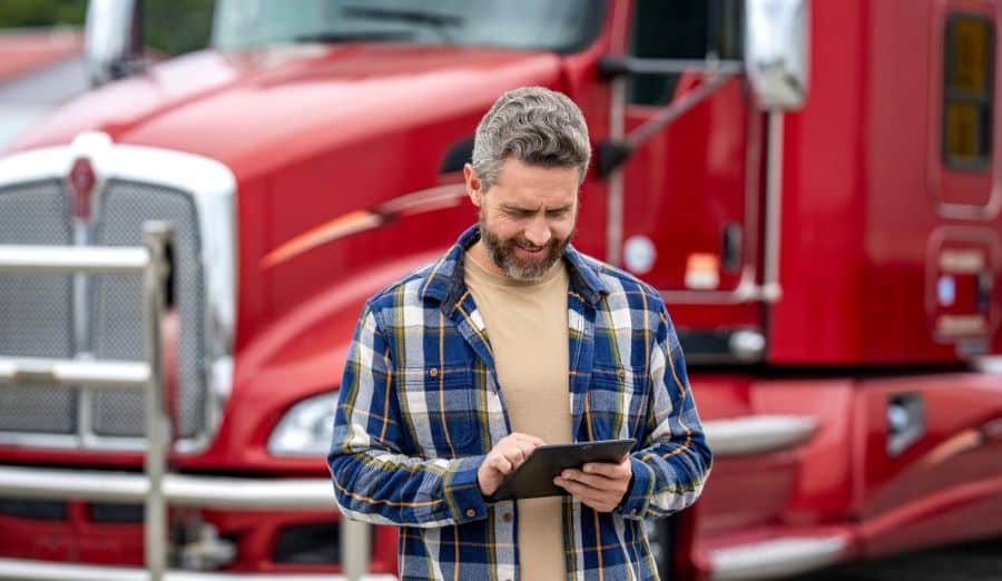 Un homme vêtu d'une chemise à carreaux se tient devant un semi-remorque rouge, souriant tandis qu'il utilise une tablette en extérieur — illustrant ainsi comment les chauffeurs peuvent facilement gérer la planification des quais et optimiser leurs opérations grâce aux technologies modernes. C3 Solutions