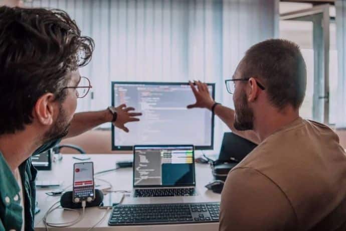 Deux collègues travaillent ensemble tout en désignant des lignes de code relatives à des systèmes de gestion d'entrepôt et de transport, affichées sur l'écran d'un ordinateur de bureau et celui d'un ordinateur portable. C3 Solutions